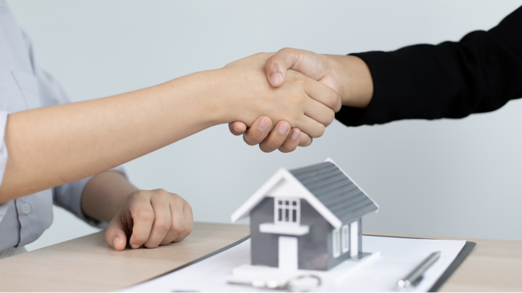 Two people shaking hands over a miniature house on a clipboard with keys nearby, symbolizing a successful real estate deal or agreement.