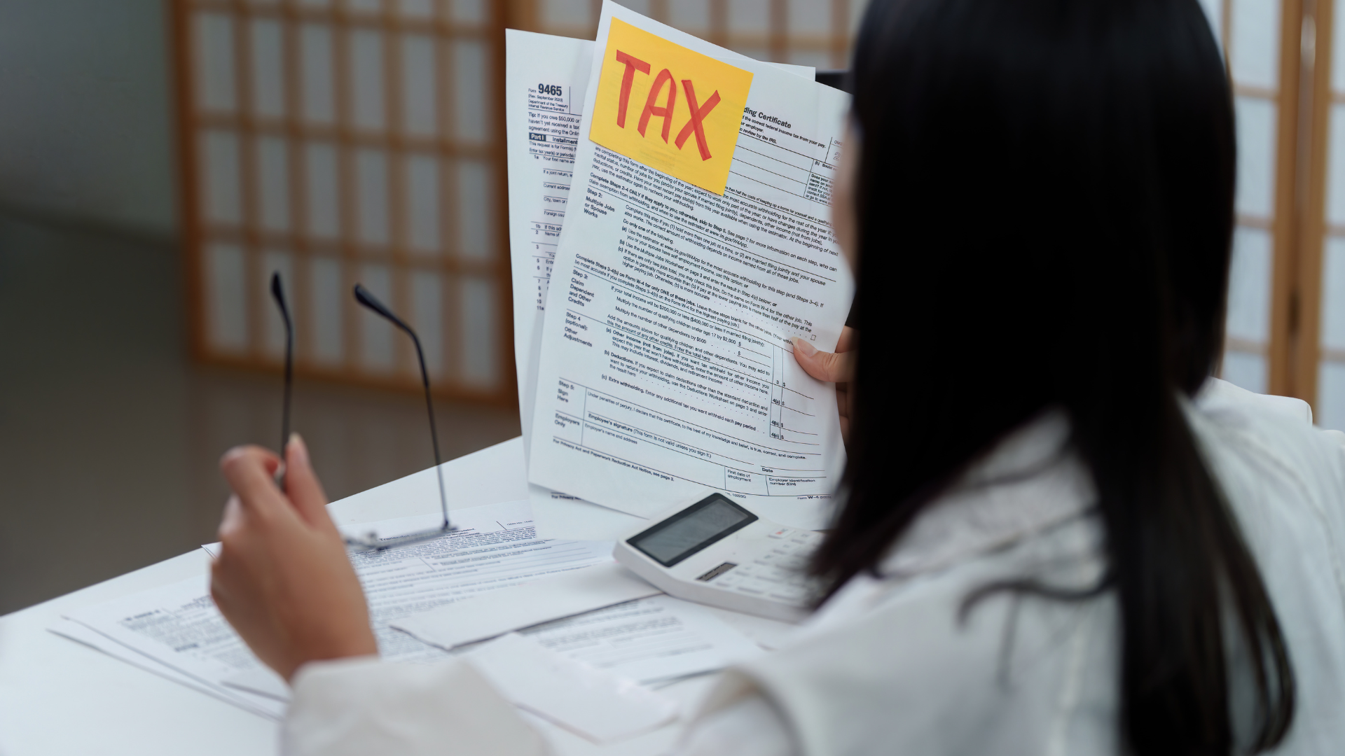 A person holding tax documents labeled "TAX" sits at a table with a calculator and glasses, conveying focus and seriousness in financial matters.