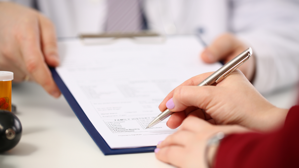 A person is filling out a medical form on a clipboard handed by a doctor. A pill bottle and stethoscope are visible, indicating a clinical setting.