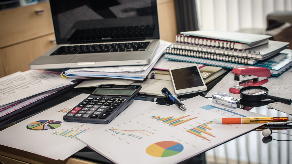 Cluttered office desk with a laptop, paperwork, charts, notebooks, calculator, and other work supplies.