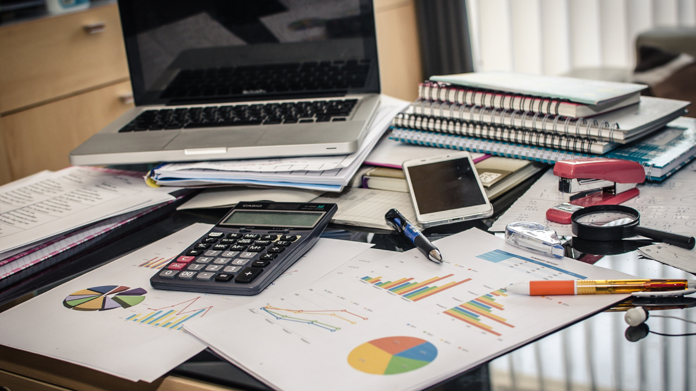 Cluttered office desk with a laptop, paperwork, charts, notebooks, calculator, and other work supplies.